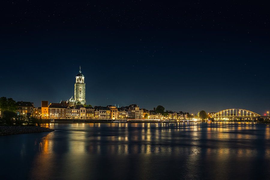 Prachtige nachtelijke foto van Deventer met de verlichte Lebuinustoren en Wilhelminabrug, weerspiegeld in de IJssel. Perfect voor liefhebbers van stadsfotografie en historische Nederlandse steden, brengt dit werk sfeer en elegantie in elk interieur.